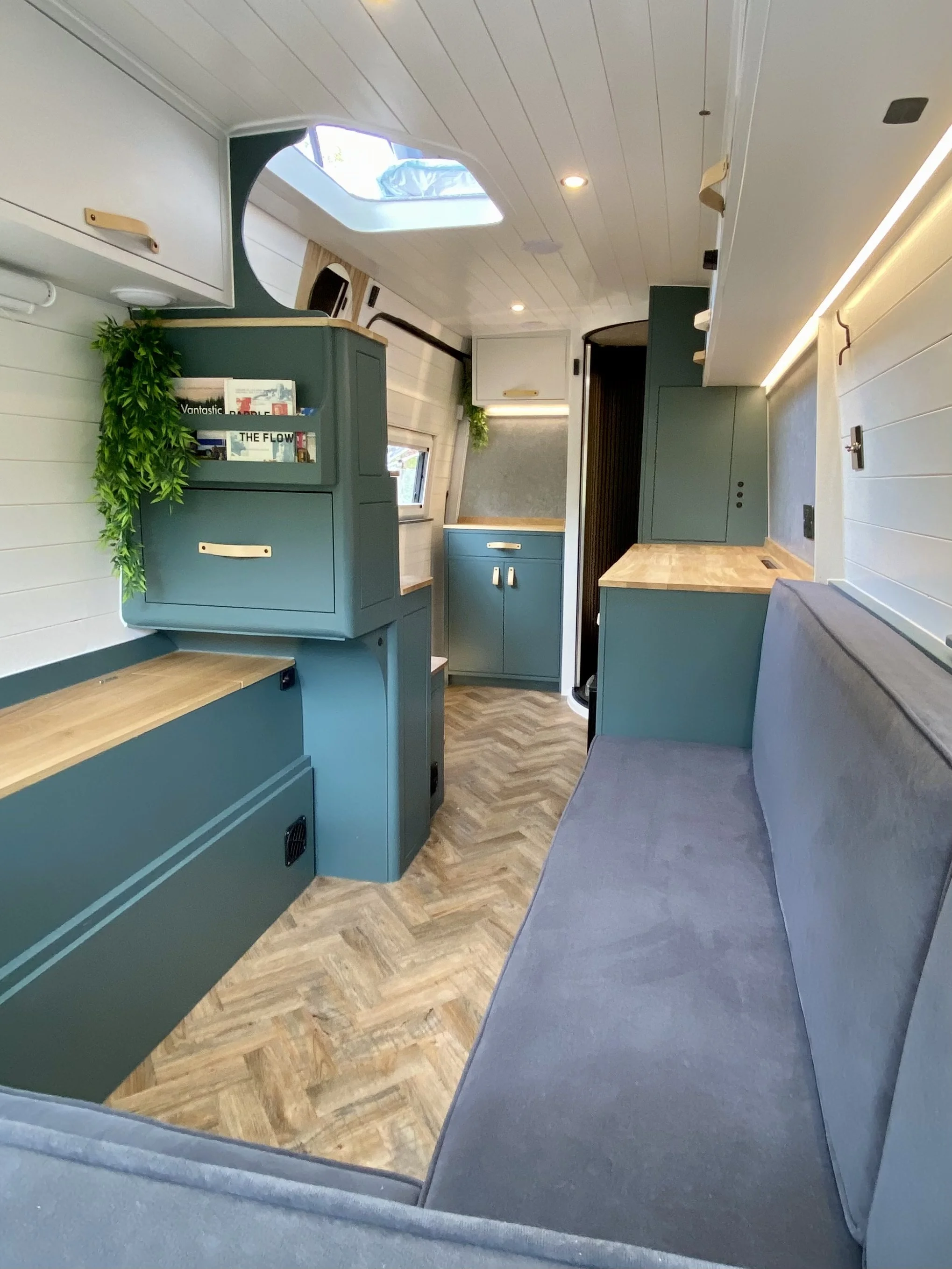 Interior of a tiny house or camper with teal cabinets, a gray cushioned seating area, wooden countertops, small windows, and a skylight in the ceiling, with wood-look flooring.