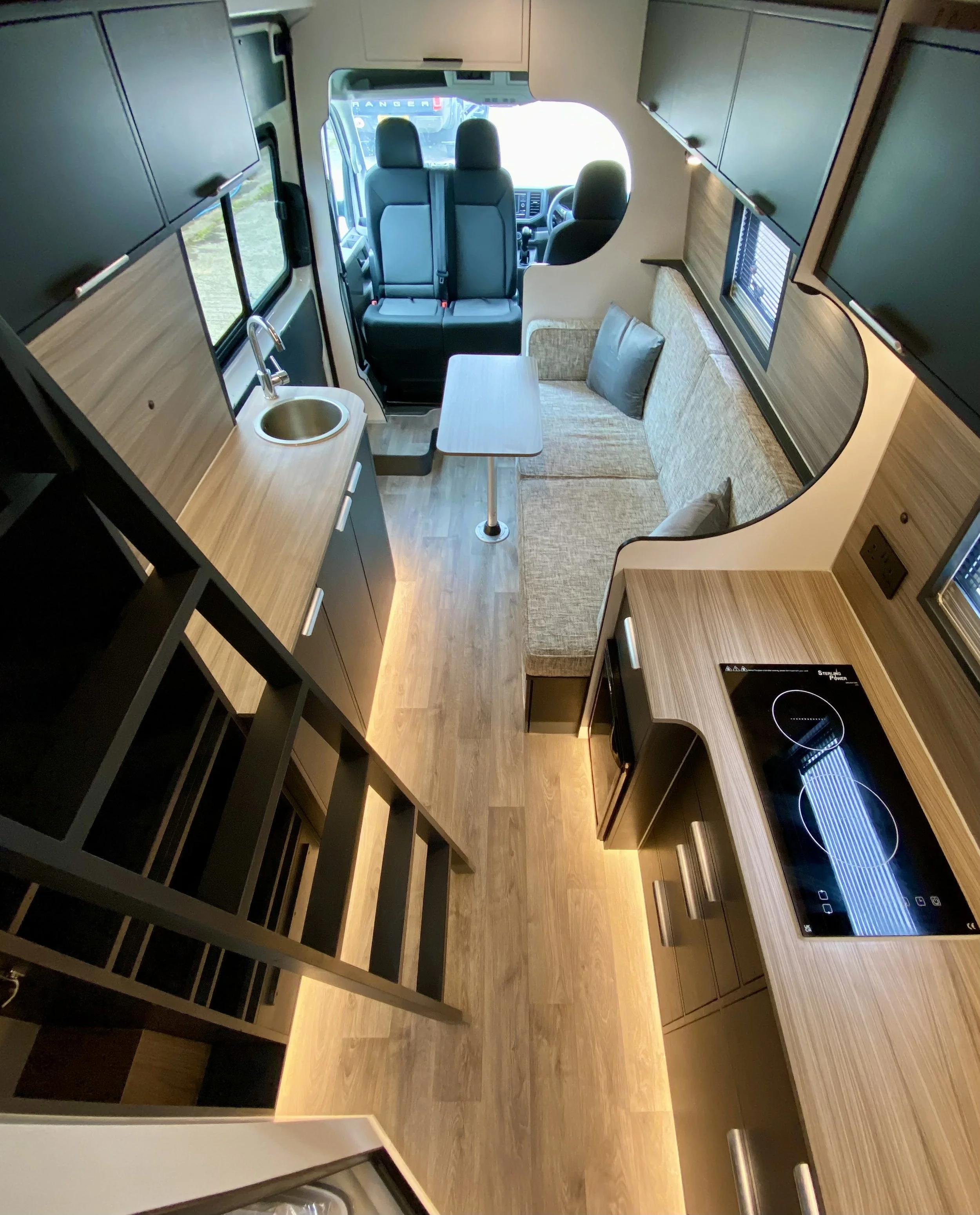 Interior view of a camper or RV showing a small kitchen area with a sink, stovetop, and cabinets, next to a seating area with a couch and cushions, with the driving cab visible through an open partition.