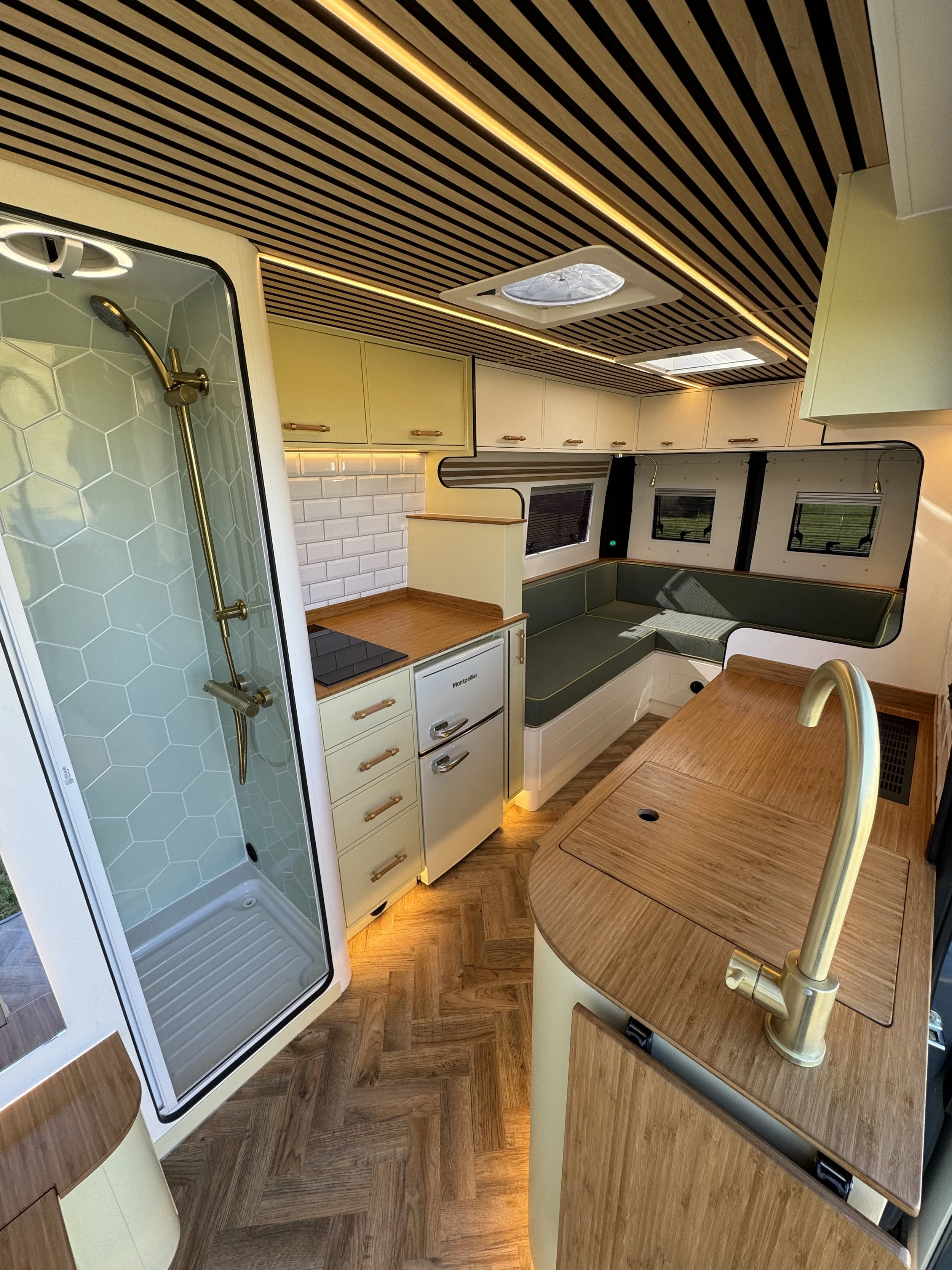 Interior of a modern camper van with a shower, kitchenette, and seating area. The ceiling is wood-paneled with integrated lighting, and there are windows with shades. The kitchenette has a stove, cabinets, and a small fridge. The seating area has a corner sofa with cushions and a wooden table.