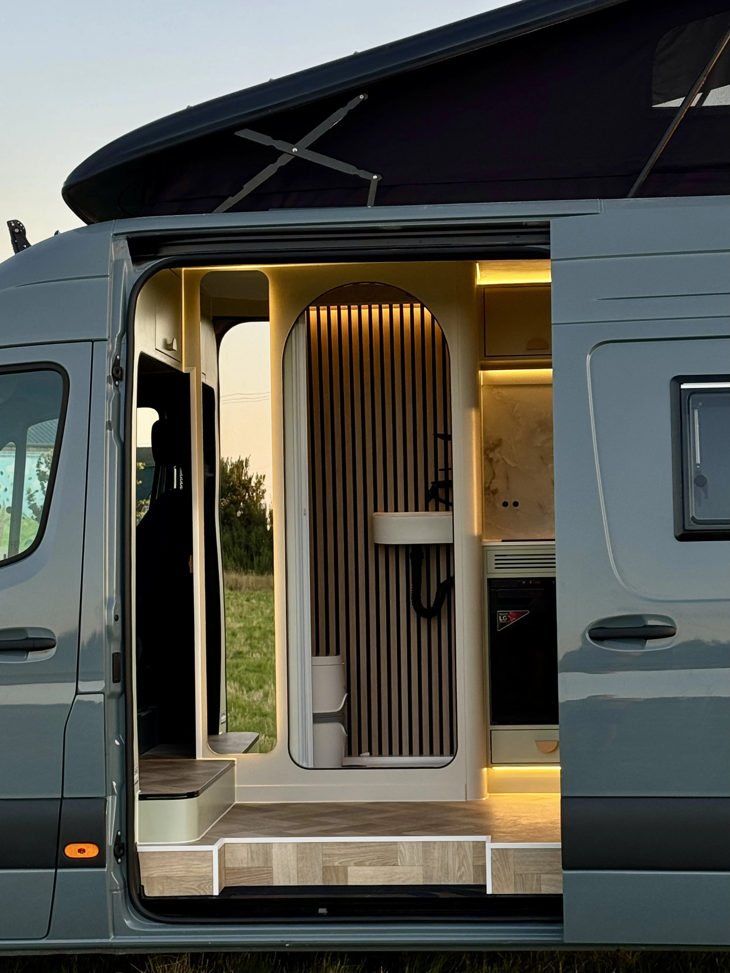 Interior of a modern camper van with wooden flooring, light-colored walls, and ambient lighting, including a small sink and a section of vertical wooden slats.