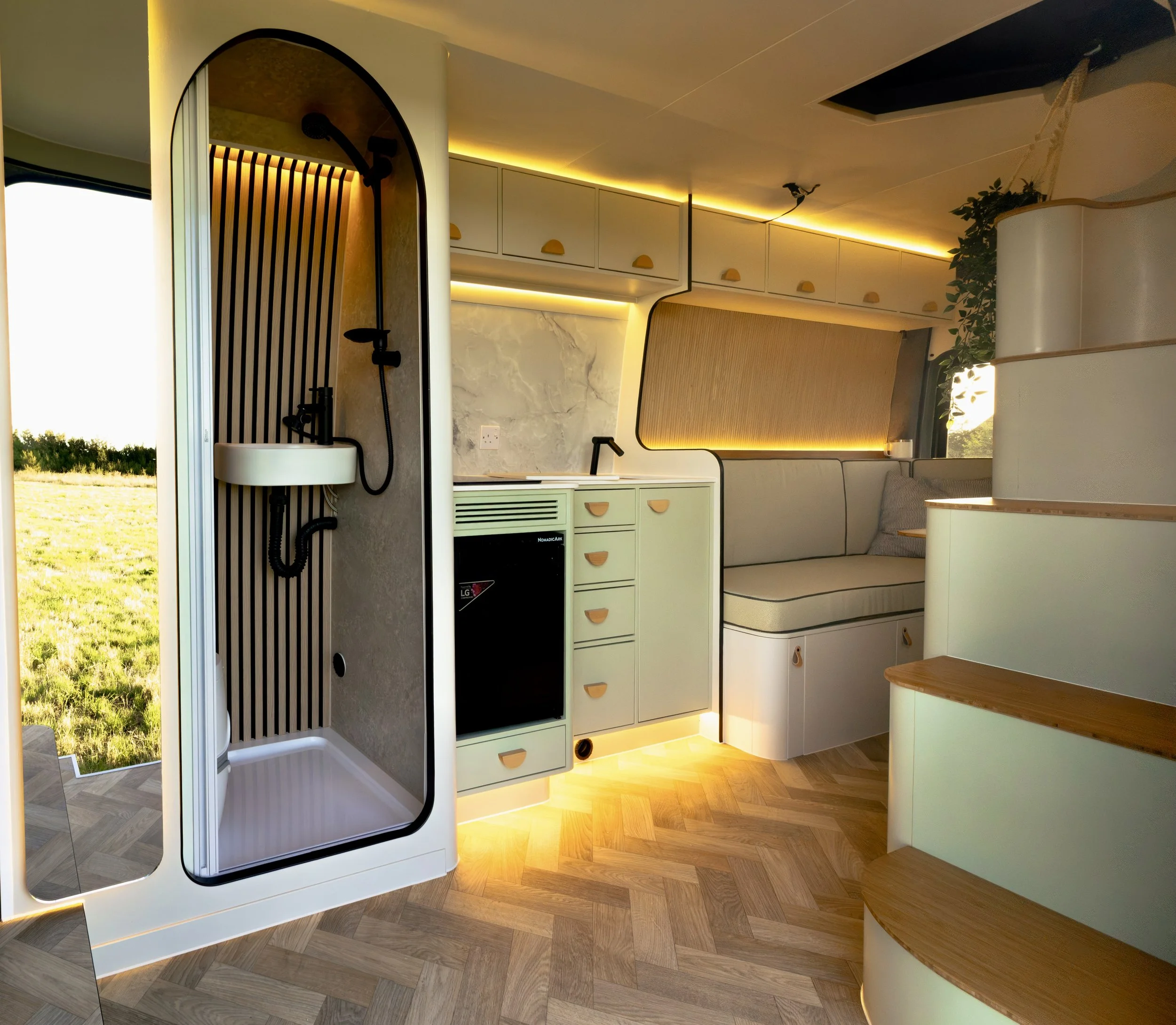 Interior of a modern tiny home with a compact kitchen, a curved corner sofa, and an outdoor shower area visible through a door, with warm lighting and wooden flooring.