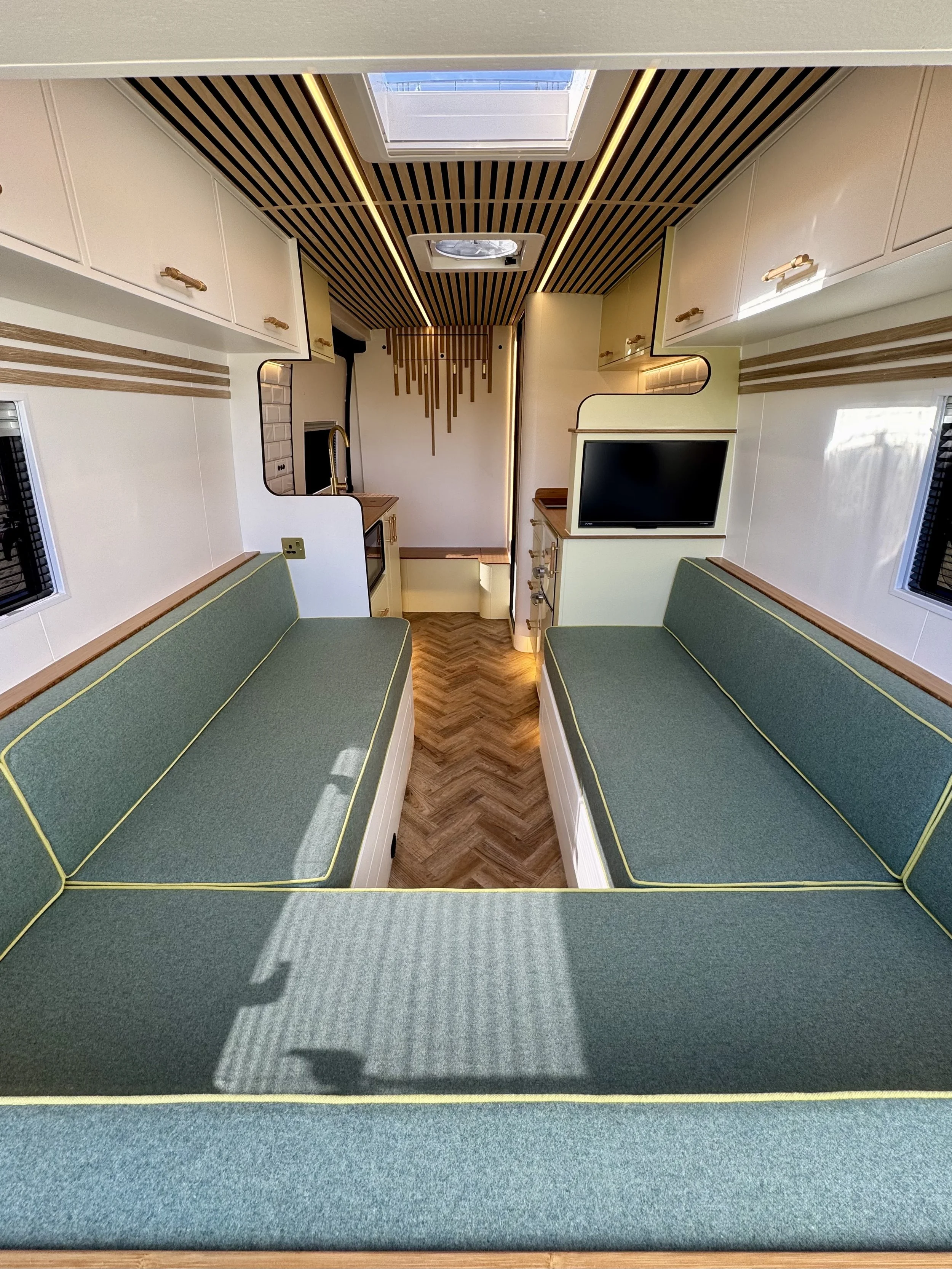 Interior of a compact living space, possibly a camper or tiny home, featuring two green upholstered benches facing each other with black and yellow piping, wooden trim, a small flat-screen TV, a kitchen area with a sink, stove, and microwave, storage cabinets, a skylight, and warm wood and white finishes.