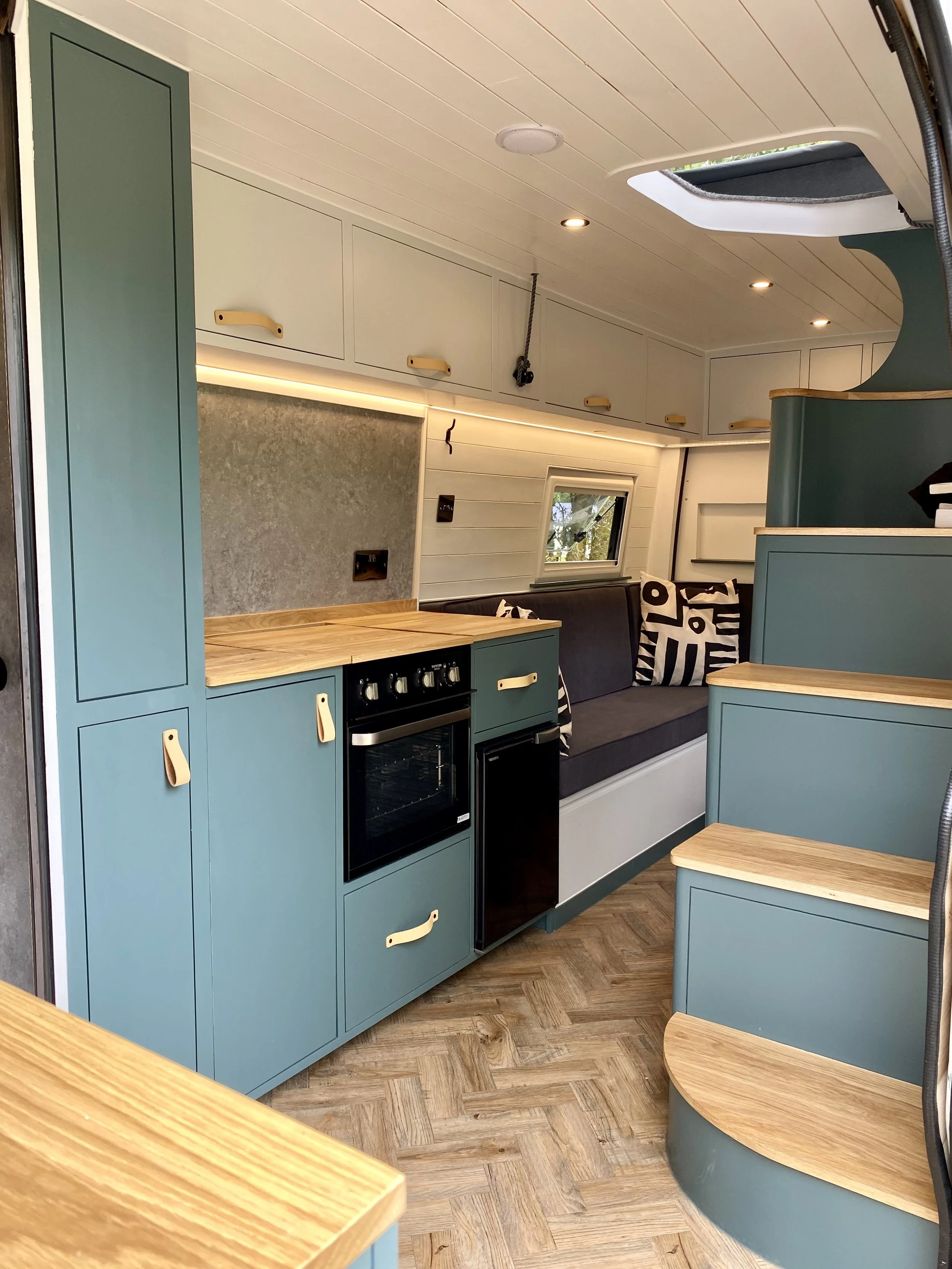 Interior of a tiny home with blue cabinetry, a black oven, light wood countertops, and a window. There is a small seating area with black and white pillows and a ceiling hatch.
