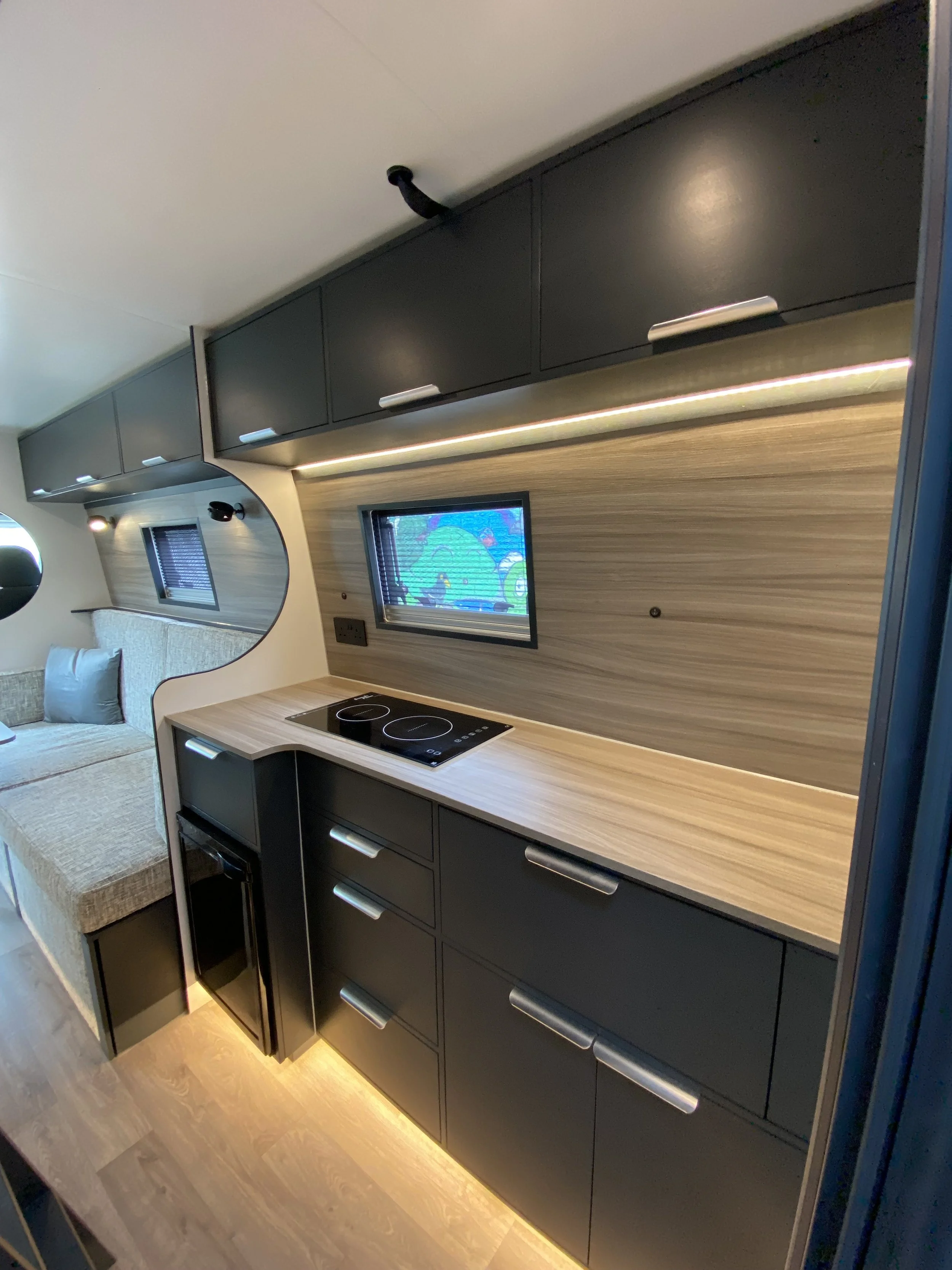 Interior of a modern RV or camper with black cabinetry, wooden countertop, a small electric cooktop, a built-in television, and a sofa with pillows.
