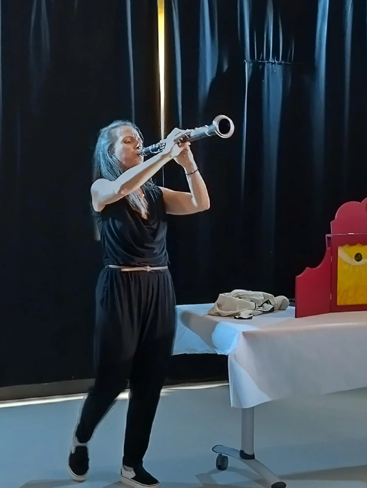 Une femme joue de la clarinette devant un décor noir, avec une table recouverte d'une nappe blanche. Elle porte un vêtement noir et des chaussures noires et blanches.