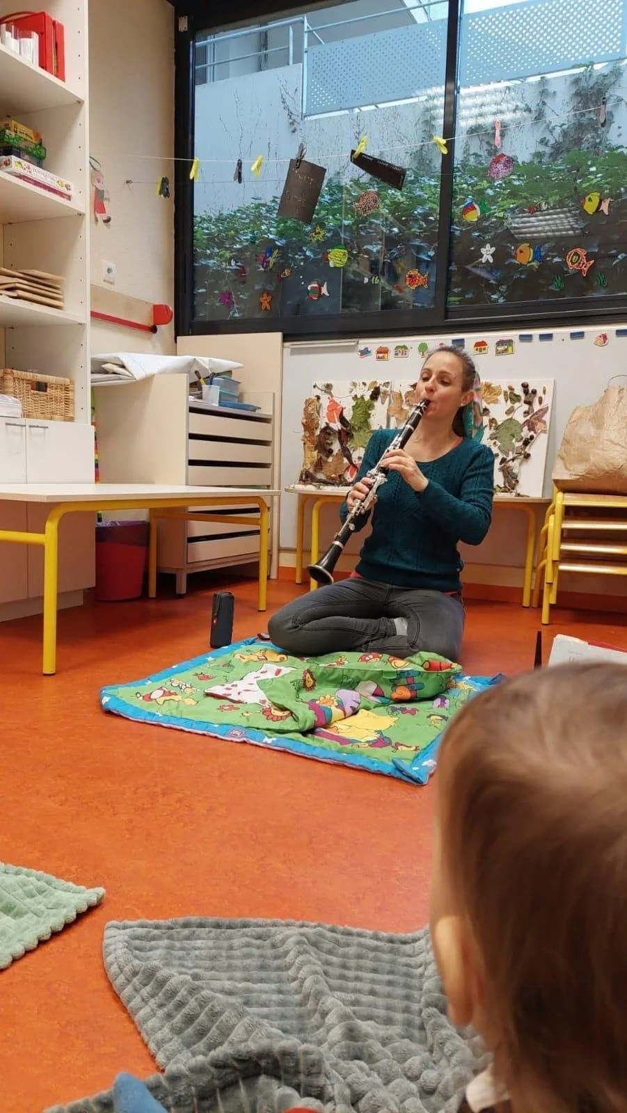 Une femme joue de la clarinette assise sur un tapis dans une salle de classe ou un espace de garde d'enfants, avec des décorations colorées et un grand tapis illustré.