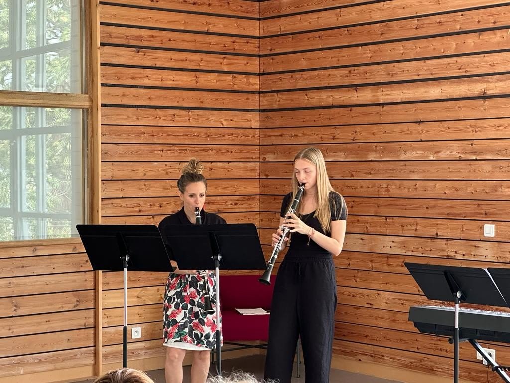Deux jeunes femmes jouent de la musique dans une pièce au mur en bois, avec une grande fenêtre à gauche et un piano noir à droite.