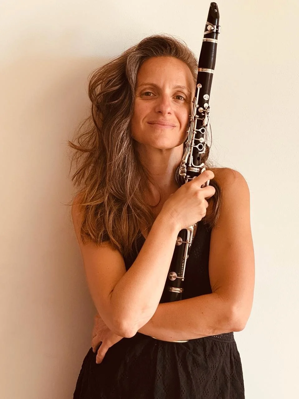 Une femme avec cheveux longs et blonds, vêtue d'une robe noire, tient un hautbois contre son visage, souriante, devant un mur blanc.