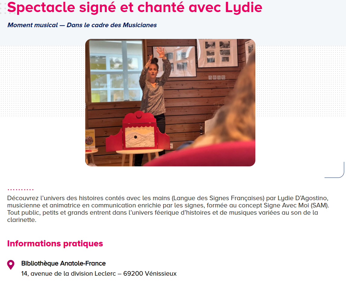 Une femme chante et mime avec ses mains lors d'un spectacle pour enfants dans une salle en bois, devant un tableau noir en décor.