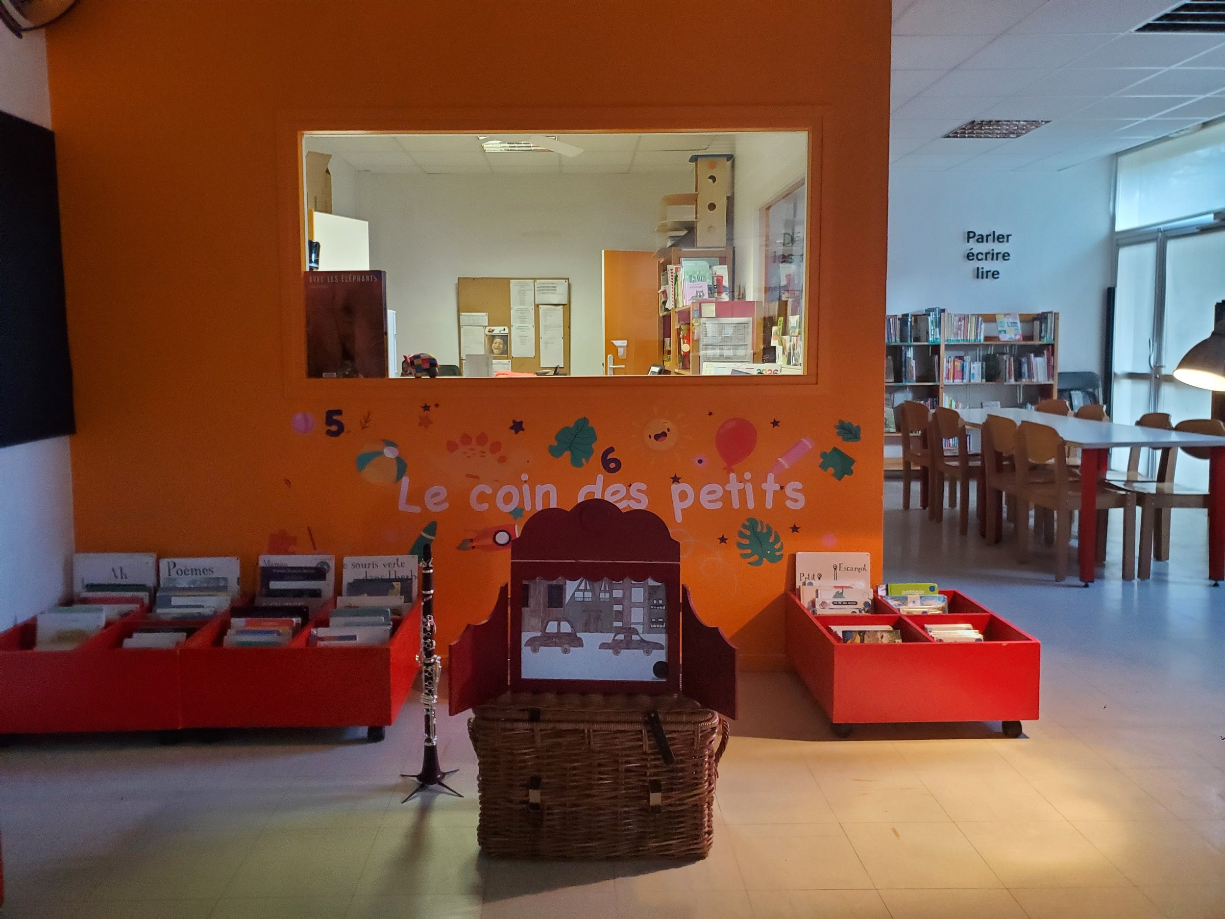 Espace de lecture pour enfants avec des étagères remplies de livres, une décoration colorée sur un mur orange avec le texte "Le coin des petits", et une vue partielle d'une salle avec des tables et des chaises.