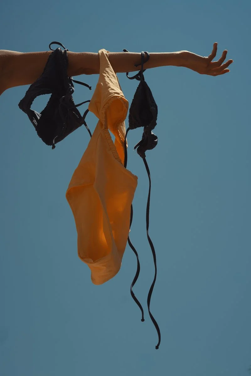 Three Arloe swim pieces hanging out from an arm, with blue sky as bakcground