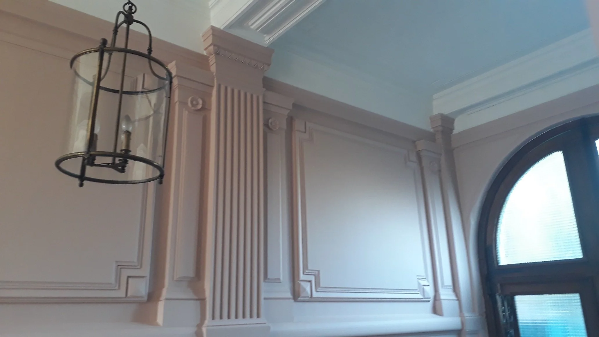 Panneau en bois peint beige avec moulures décoratives, partie d'une salle élégante ayant une grande fenêtre avec volets et un lustre noir suspendu.