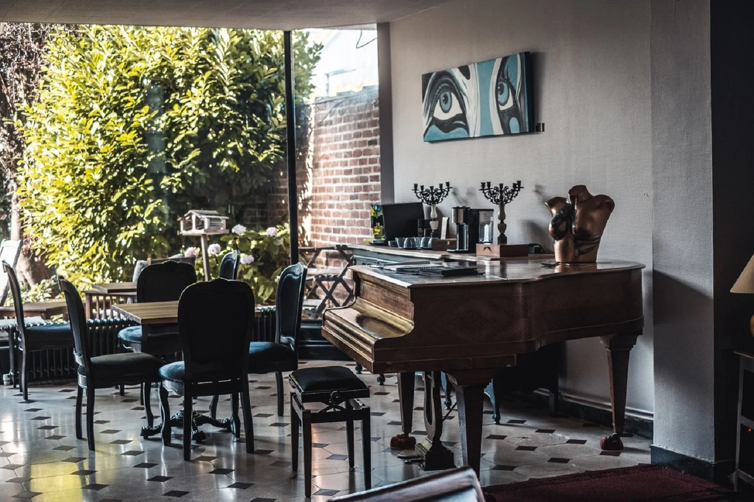 Intérieur d'une salle à manger avec une grande fenêtre donnant sur un jardin vert, un piano en bois, une peinture abstraite au mur, deux candélabres, un ordinateur et une machine à café sur une table à côté du piano.