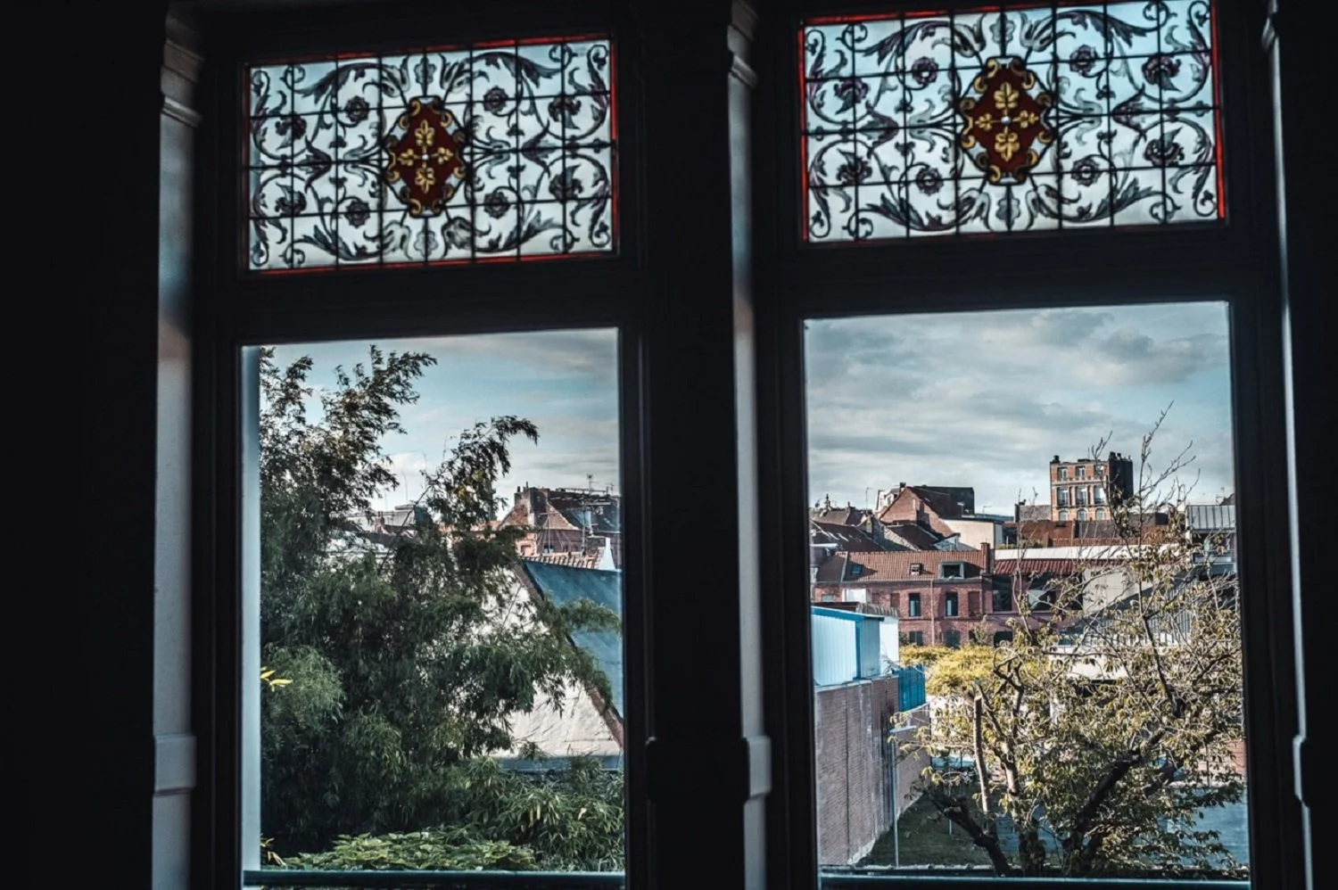 Vue d'une fenêtre en vitre avec des vitraux décoratifs en haut, laissant voir une scène urbaine avec des bâtiments anciens, des arbres et un ciel partiellement nuageux.