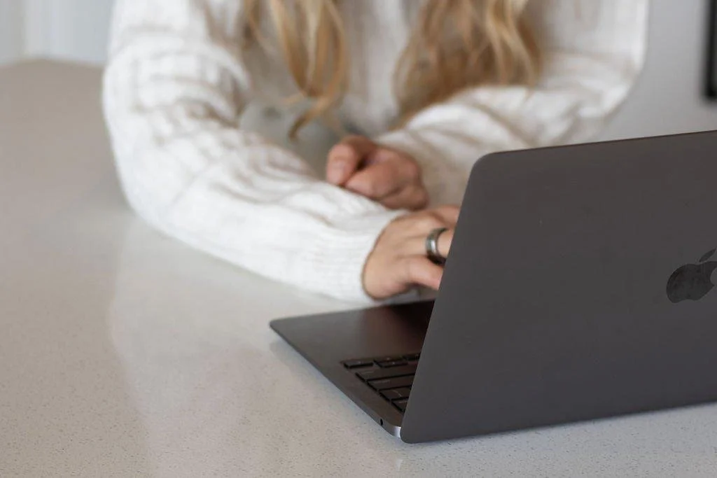 A person with long blonde hair wearing a white sweater sitting at a desk with a black MacBook laptop in front of them, arms crossed, on a light-colored surface in a bright room.