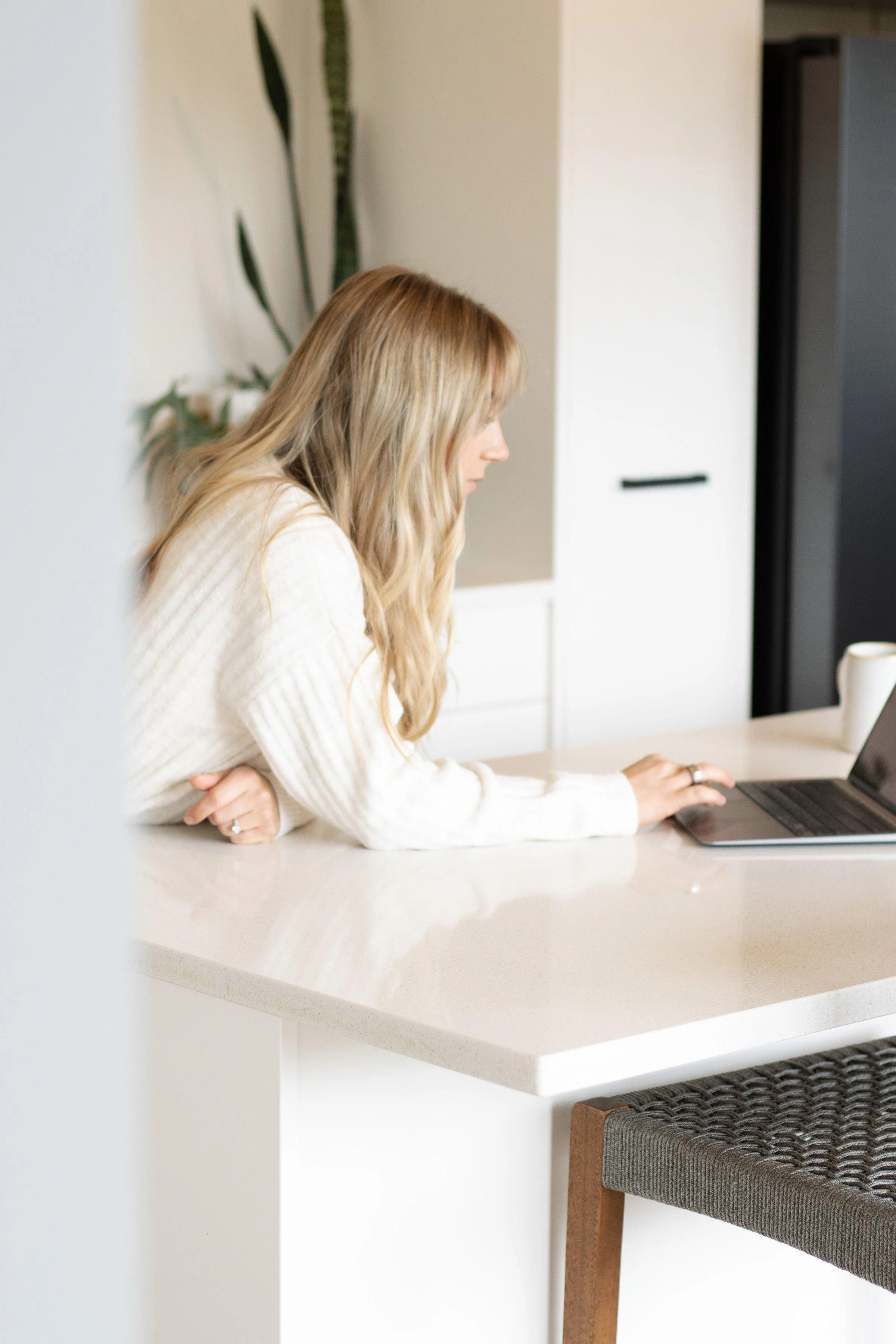 A woman with long blonde hair sitting at a white kitchen counter working on a laptop.