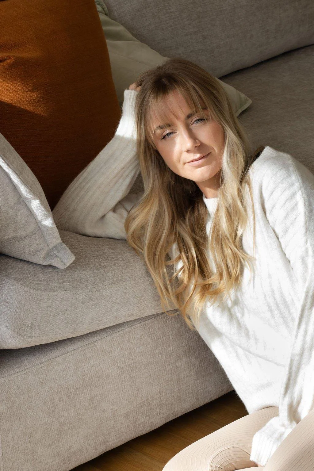 A woman with long blonde hair, wearing a white knit sweater, is lying on a beige sofa with brown and beige pillows, looking at the camera with a relaxed expression.