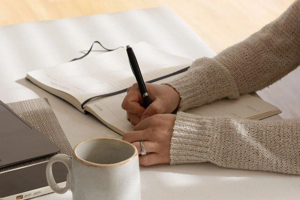 A person wearing a beige sweater writing in a lined notebook with a black pen, on a white table, with a white ceramic mug, a closed laptop, a smartphone, and a brown placemat nearby.