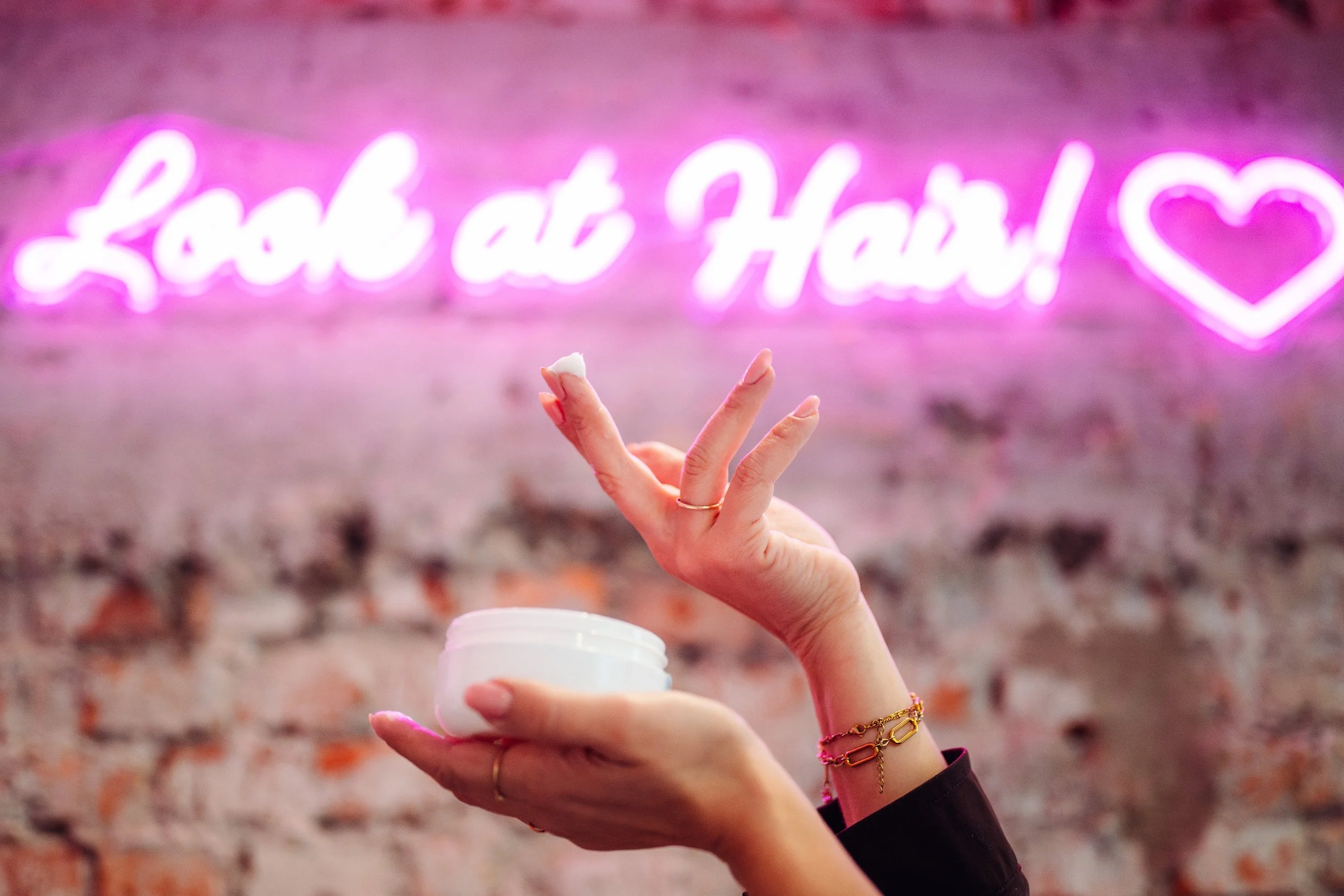 Een hand die een potje vasthoudt met een witte vloeistof en een vinger die wordt uitgestoken, met een achtergrond van een roze neonlichttekst dat zegt "Love at Heart" met een hartvorm.