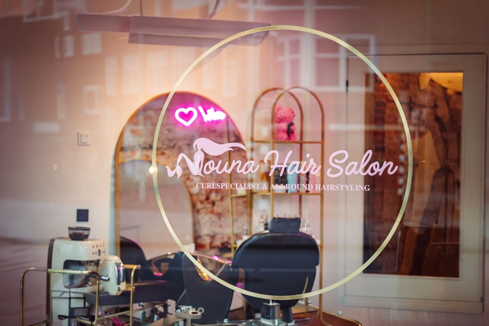 Raam van een kapsalon met decoratie, een spiegel, een stylingstoel, en een kopieermachine in een winkel met de naam 'Mouna Hair Salon'.