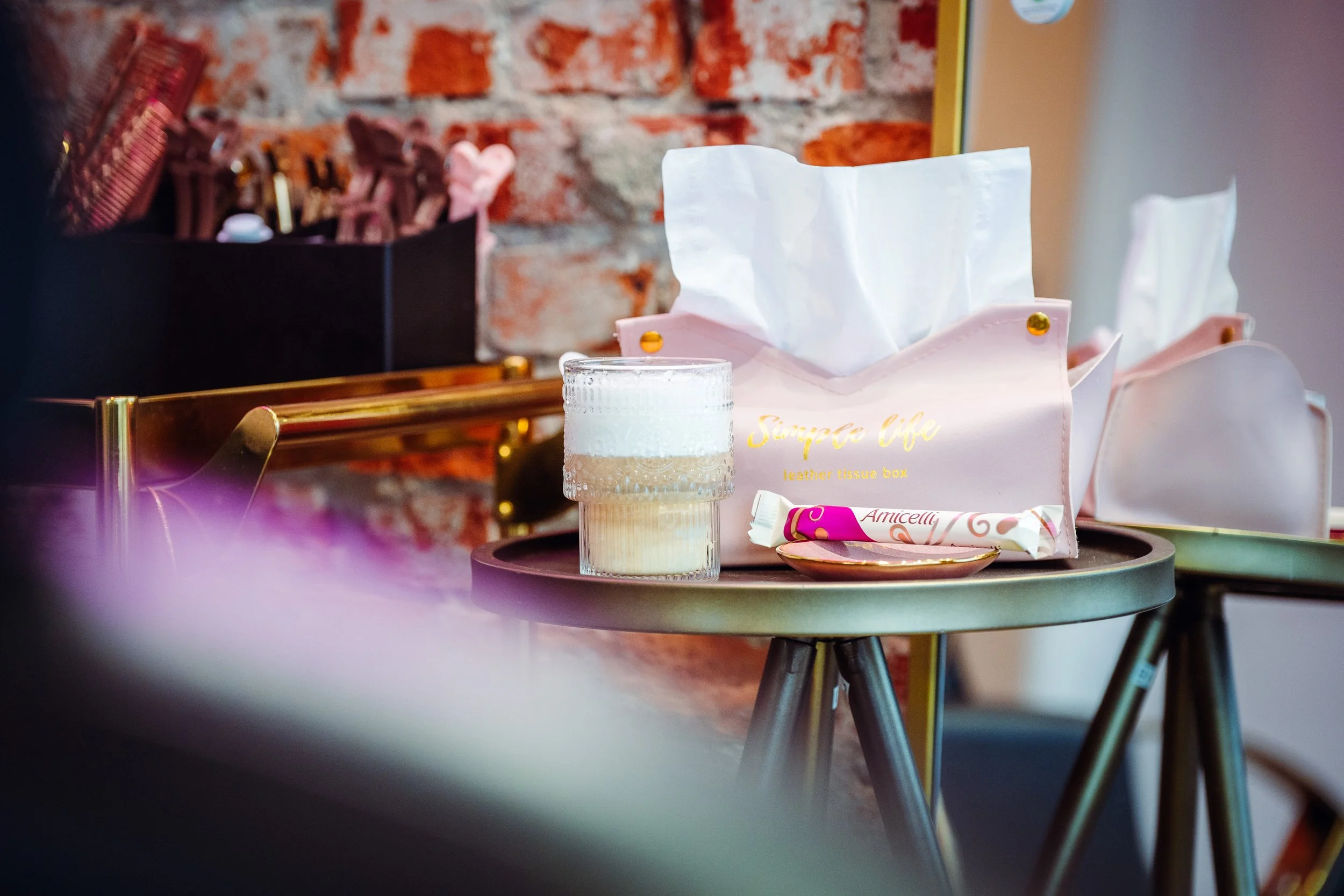 Een tafel met een glas koffie, een roze zakje met de tekst 'Serpico cafe' en andere kleine voorwerpen, tegen een bakstenen muur in een interieur. Naast de tafel staat een zwarte opbergdoos met haarspeldjes.