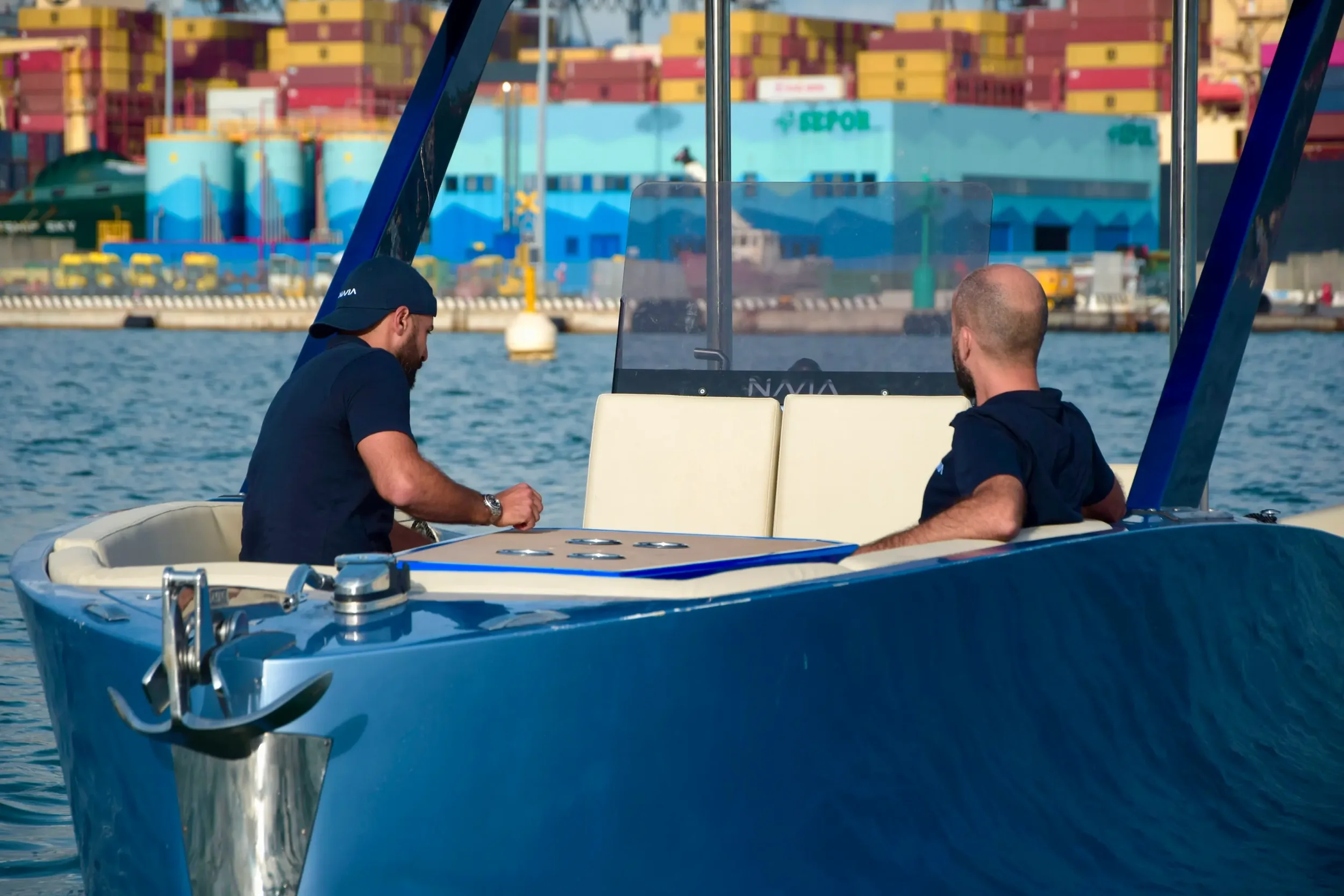 Due uomini seduti su una barca a motore, uno con cappello nero e l'altro con capelli rasati, di fronte a un porto con container colorati e acqua.