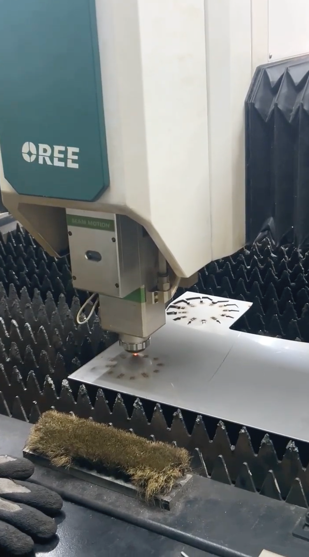 A laser cutting machine in operation cutting patterns on a metal sheet, with a robotic arm and safety guards surrounding the workspace.