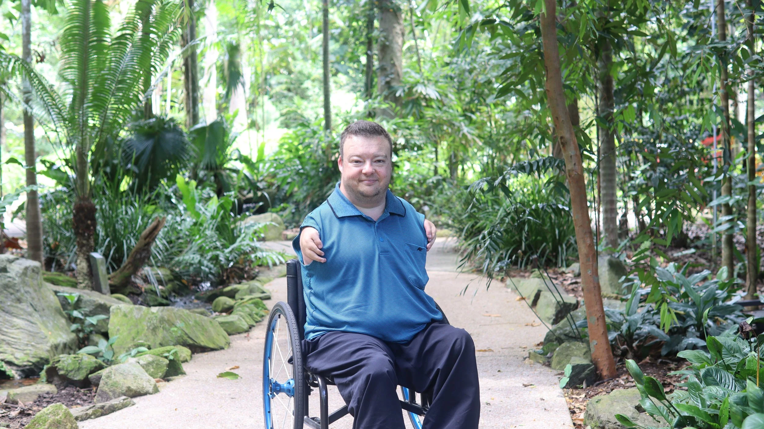 Allan Bunyan A man in a wheelchair sitting on a trail in a lush, green botanical garden. He is smiling and wearing a blue collared shirt, surrounded by various tropical plants and trees.