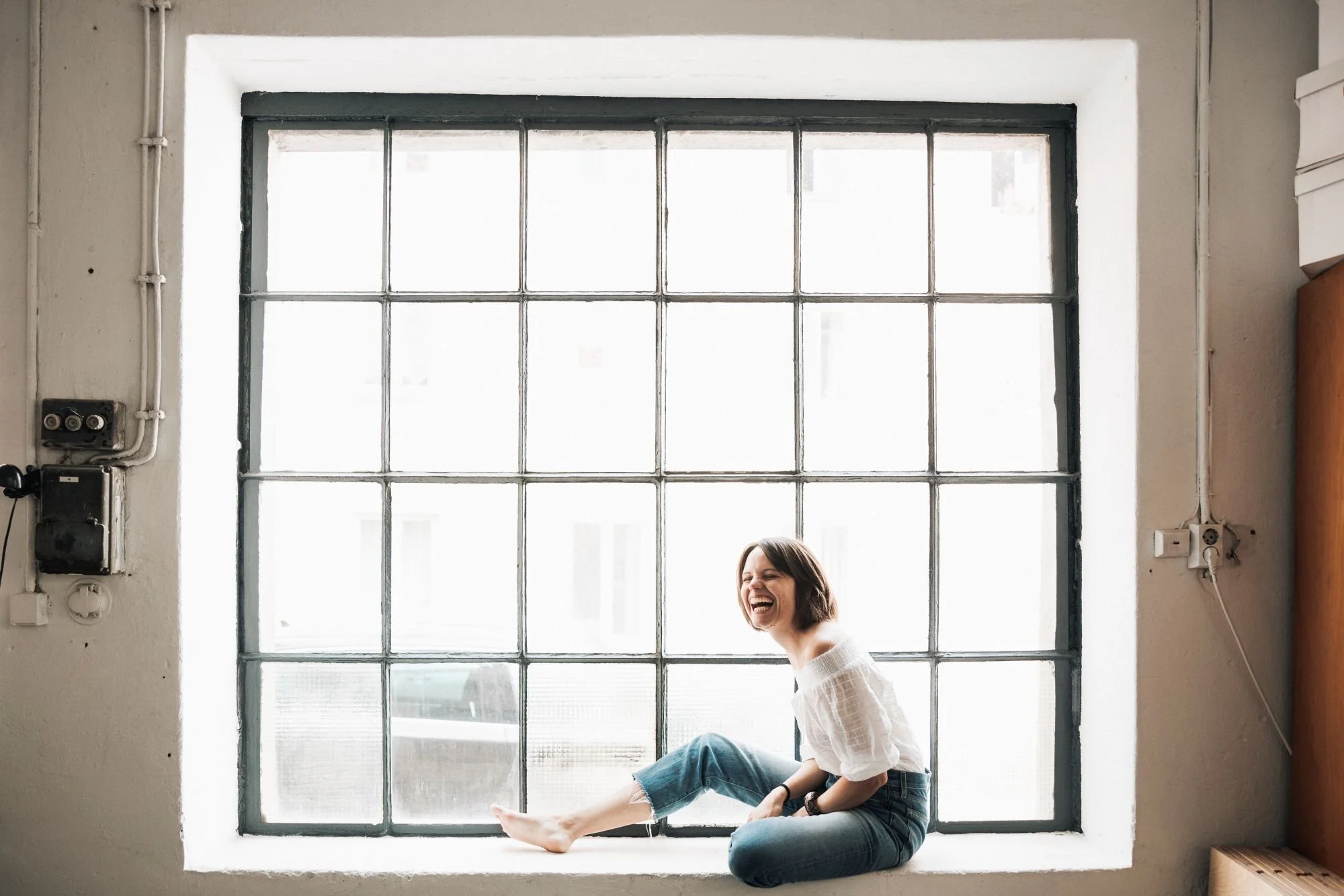 Porträtfoto von Viola Petsch, die lachend und strahlend auf einem Fenstersims vor einem industriellen Fenster sitzt.