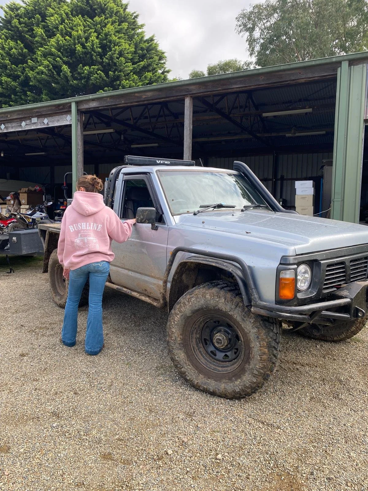 A woman in a pink hoodie and blue jeans standing next to a silver off-road Nissan patrol vehicle with large tires, mud splashes, and a black bumper.