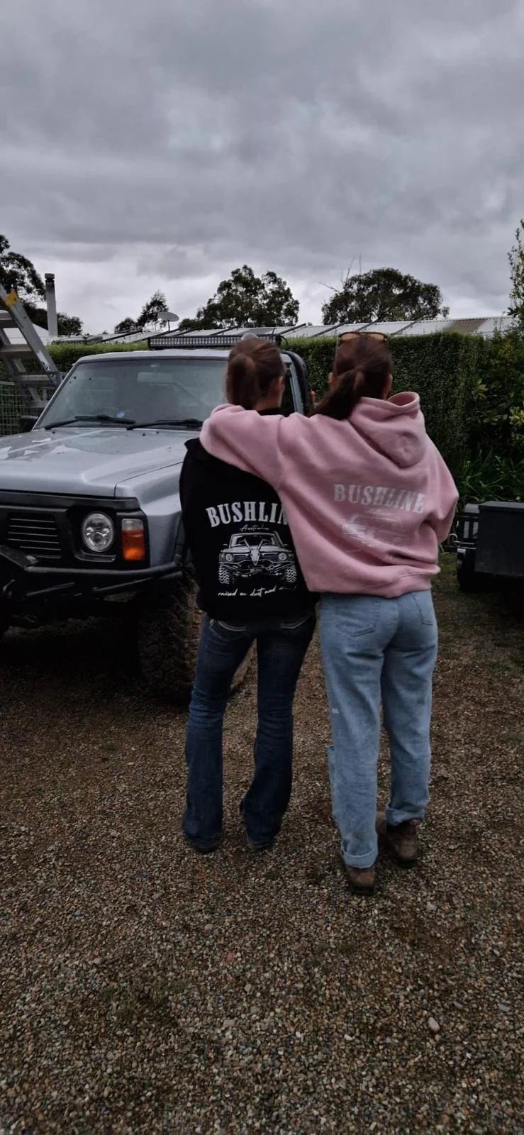 Two women standing outdoors on a gravel surface, with their arms around each other's shoulders, facing away from the camera. They are wearing hoodies in the colours black and pink as well as jeans, with a vehicle and cloudy sky in the background.