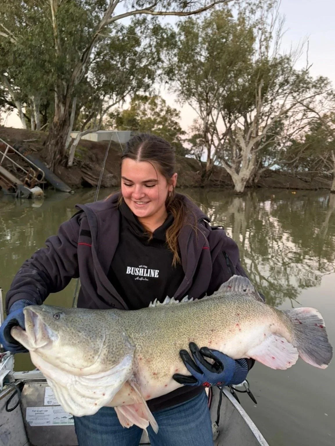 A smiling woman in a black jacket and Bushline black hoodie holding a large Murray cod fish with a water and tree background.