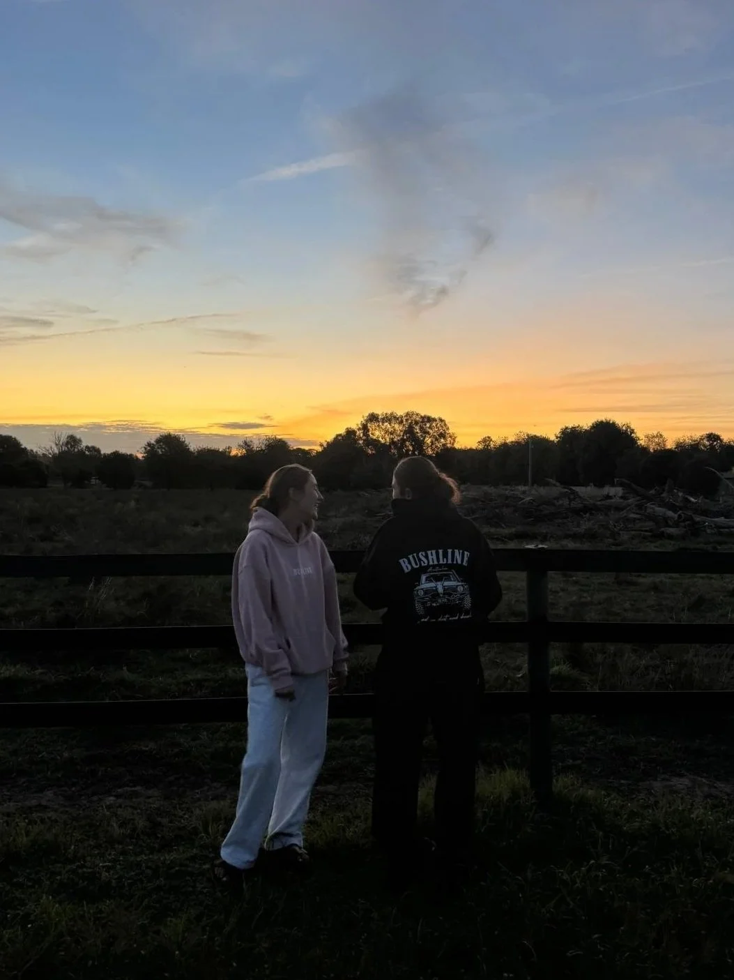 Two women standing outdoors near a wooden fence during sunset, with trees and a mostly clear sky with some clouds in the background.