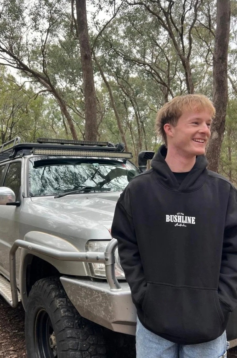 A young man with light brown hair and a black hoodie that says "BUSHLINE" is standing outdoors in front of a gray off-road vehicle with a roof rack in a wooded area.