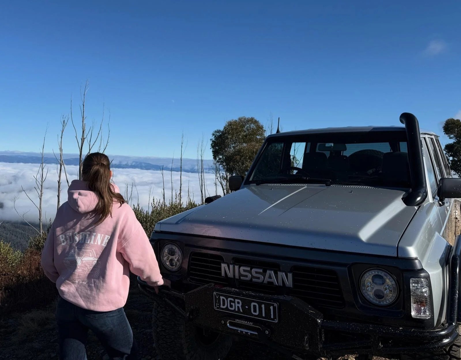 A person in a pink hoodie standing next to a silver Nissan off-road vehicle on a mountain with a view of clouds and distant mountains.