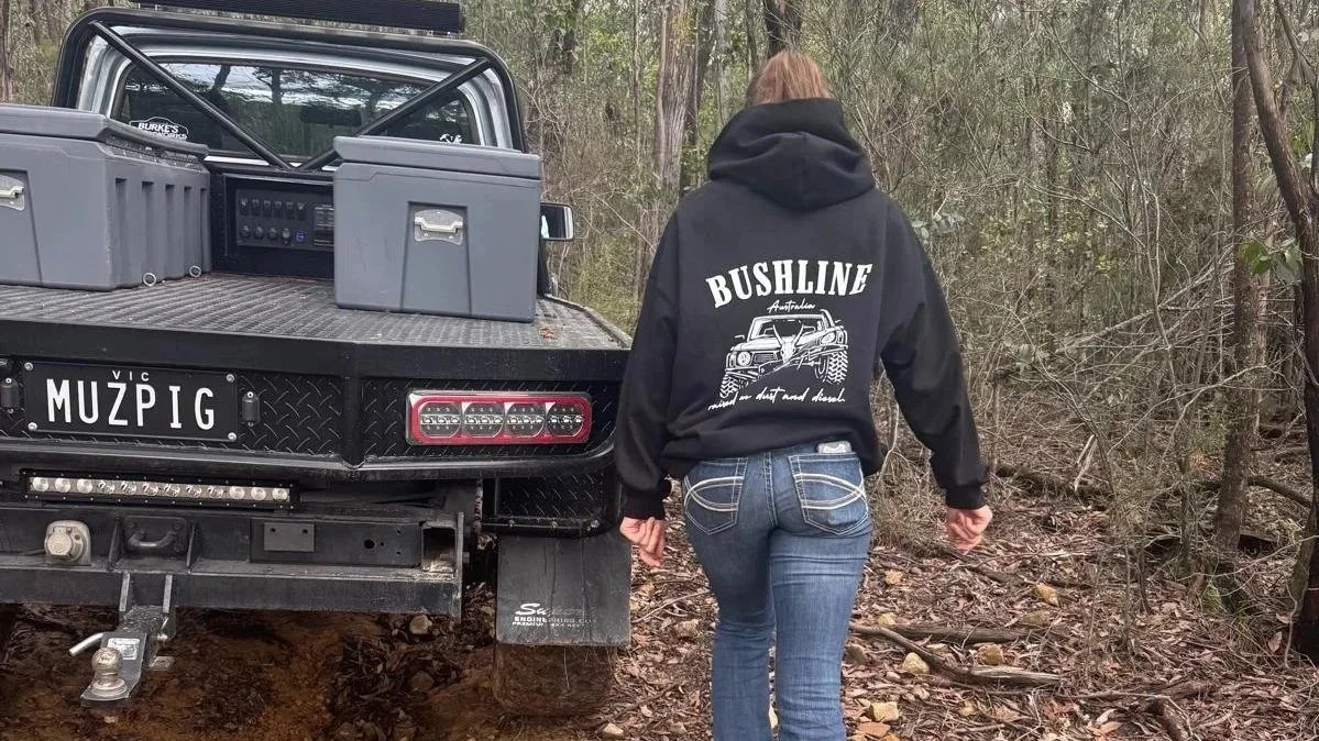 Person walking away in a wooded area, wearing a black hoodie with a graphic of a 4WD and the words 'Bushline' on the back, next to a black off-road vehicle with license plate 'MUZPIG'.