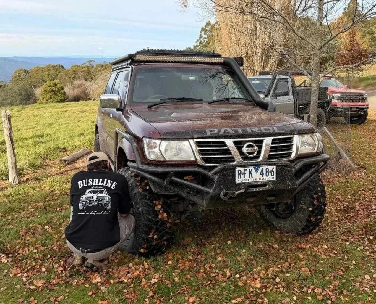A man wearing a black Bushline Australia tshirt kneels beside a black Nissan patrol off-road vehicle with large tires, a labeled front bumper, and a light bar in a grassy field surrounded by trees and hills, with other trucks parked behind.