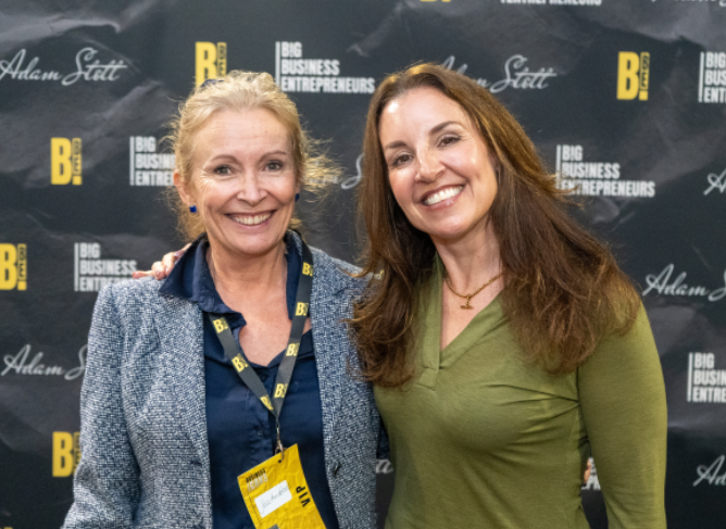 Two women smiling at a professional event, standing in front of a black backdrop with yellow and white text and logos, including "Big Business Entrepreneurs" and "Adam Statt."
