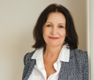 Portrait of a smiling woman with shoulder-length dark hair, wearing a white blouse and a grey blazer, standing indoors near a beige wall.