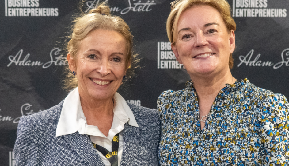 Two women smiling and posing for a photo at a business event with a black backdrop featuring the words 'Big Business Entrepreneurs' and 'Adam Stott'.