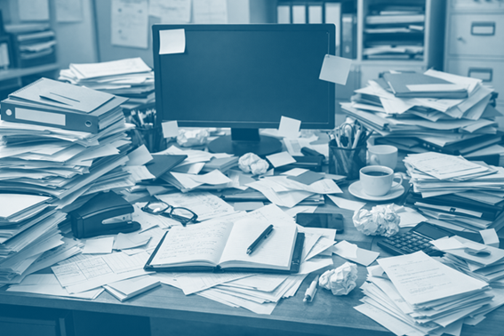 Cluttered office desk with piles of paper, a computer monitor, glasses, a notebook, crumpled paper, a coffee cup, and office supplies.