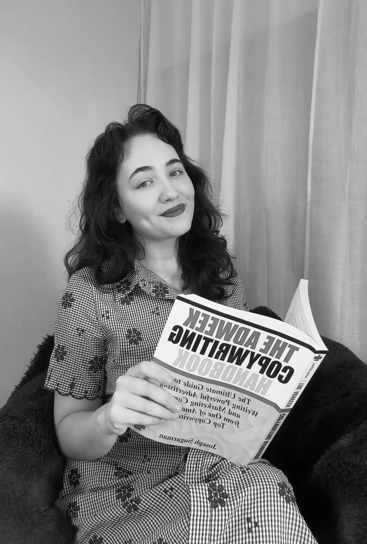 A professional photo of a confident woman with dark, curly hair, dressed in smart, polished attire, seated comfortably in a clean, modern setting—holding a notebook or laptop to reflect strategy, focus, and SEO-driven content work.