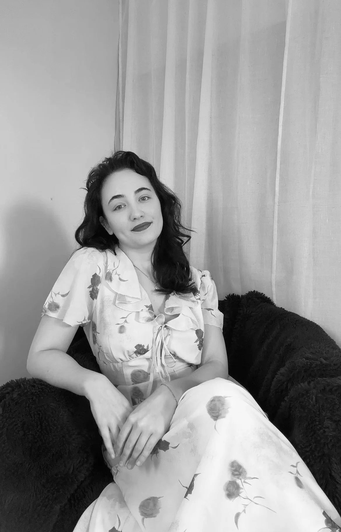 A professional image of a woman with dark, curly hair, dressed in clean, modern business-casual attire, seated on a stylish sofa in a bright, minimal setting—designed to reflect clarity, professionalism, and a calm, strategic approach to SEO content.