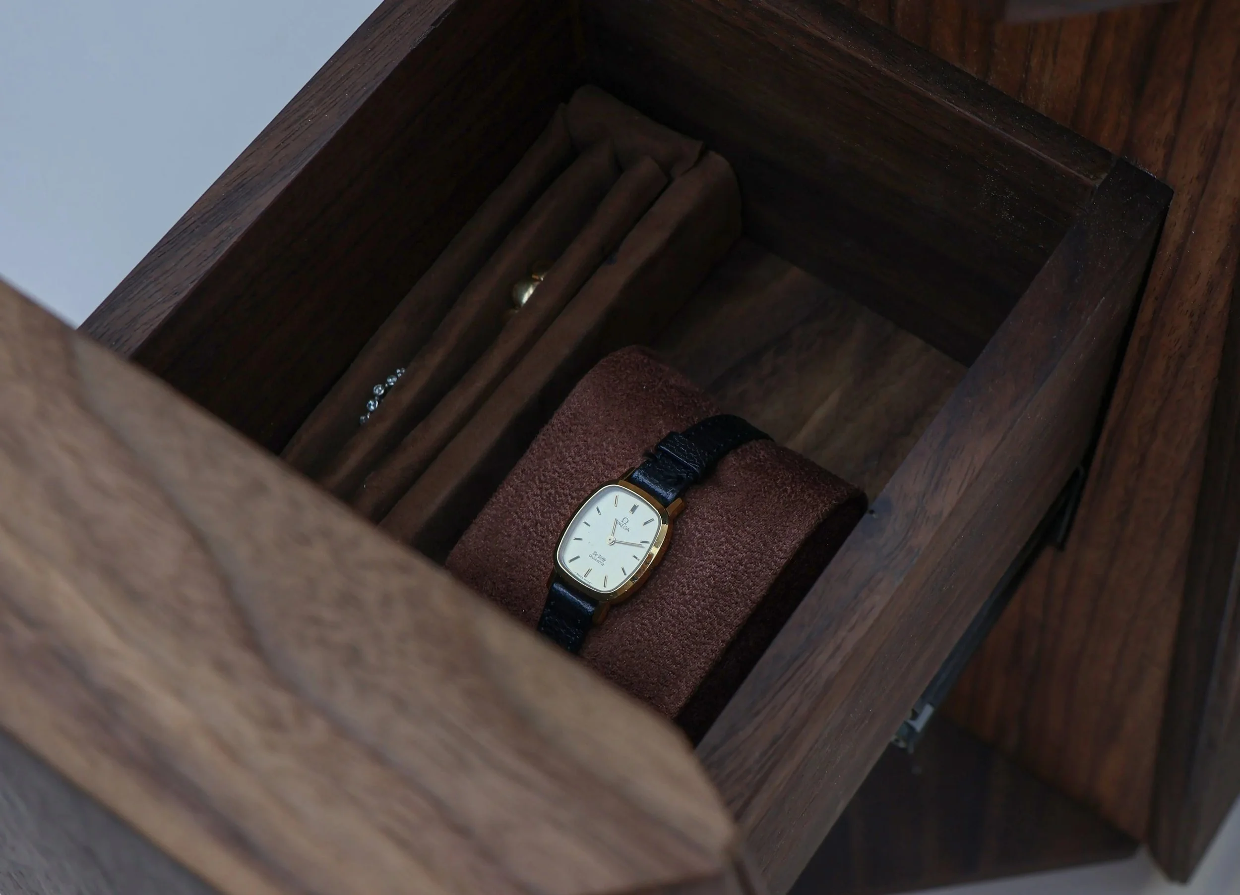 Close-up view of a wristwatch with a gold case, white dial, and black leather strap, resting inside a wooden hidden drawer with jewelry and a burgundy fabric item.