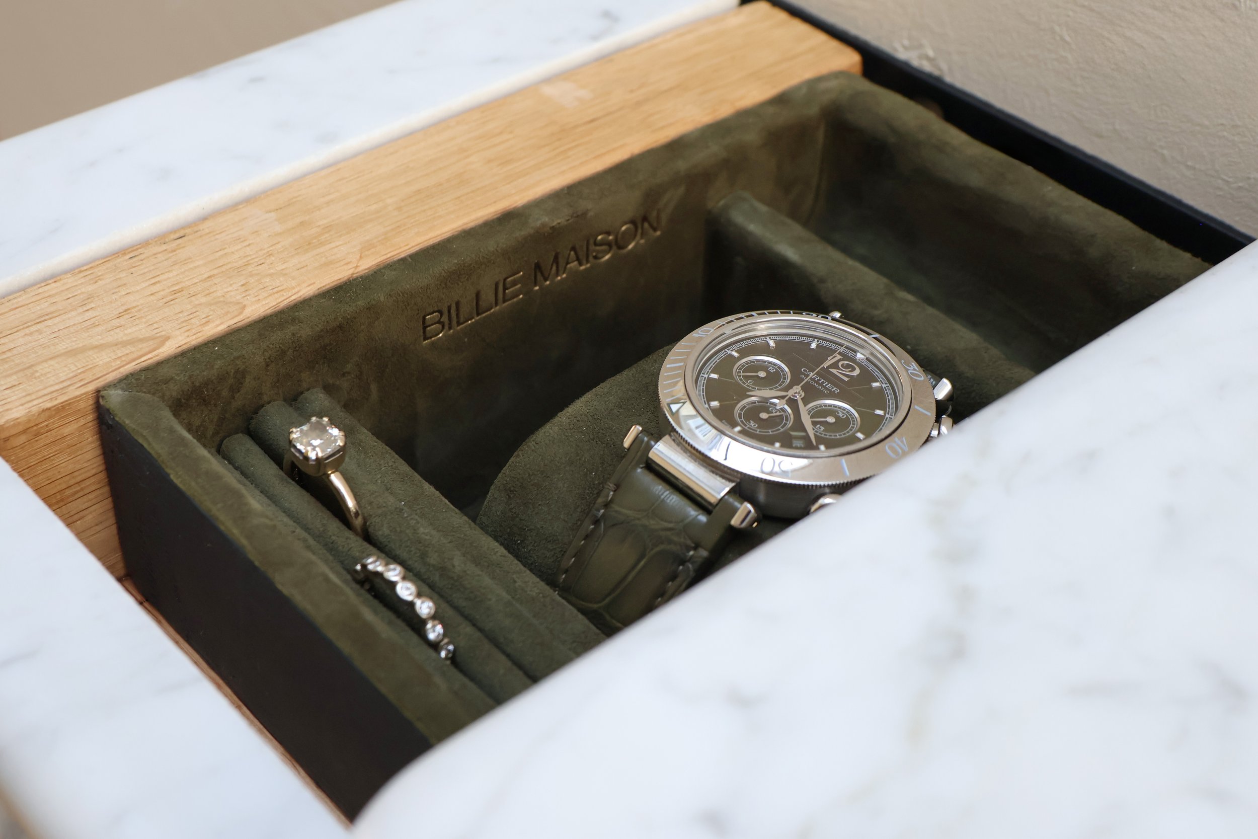 A close-up of a luxury wristwatch placed inside a dark green velvety watch box. The watch has a metallic band, a black dial with multiple smaller dials, and a rotating bezel with marked increments. The inside of the box lid has a leather patch with t