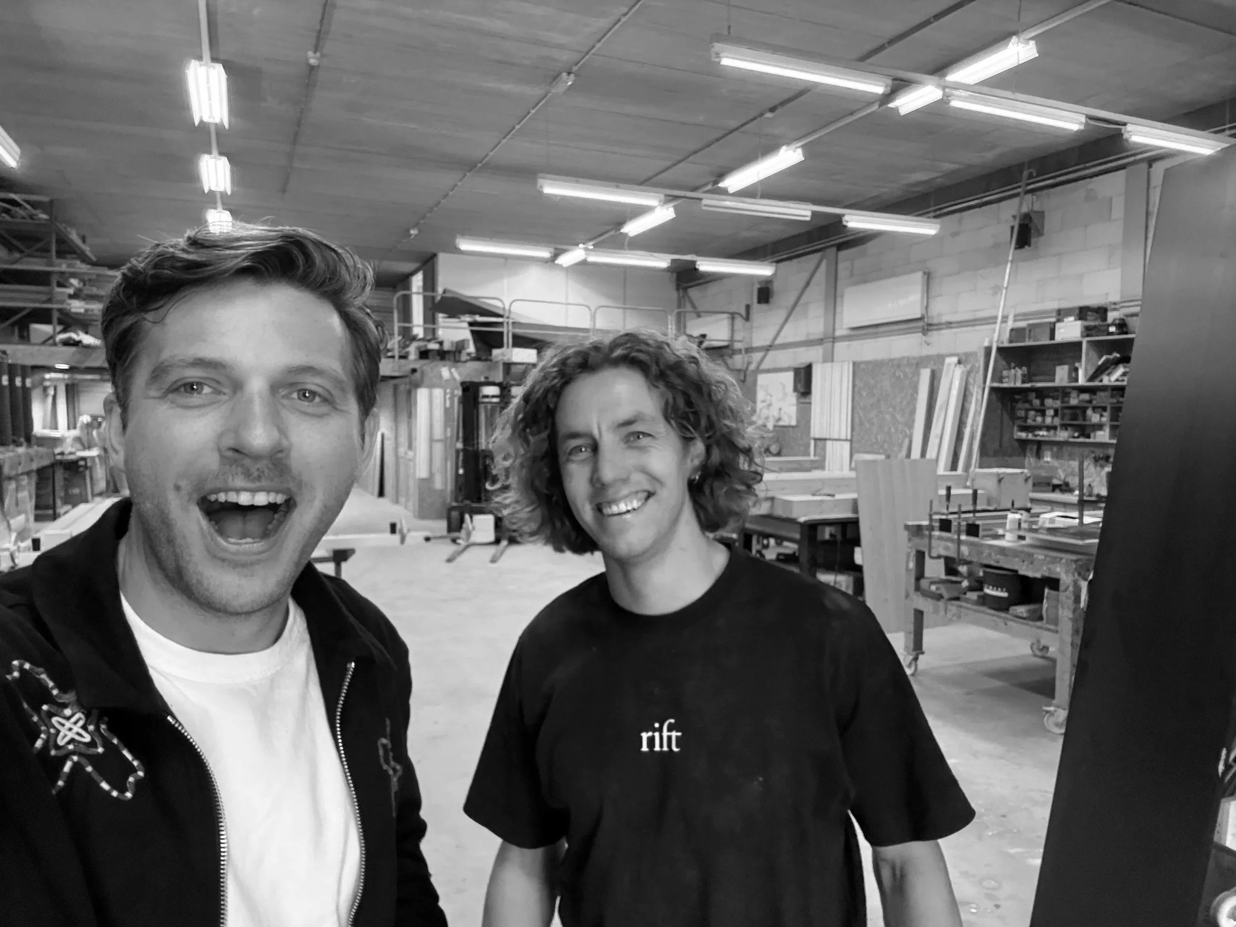 Two smiling young men taking a selfie in a woodworking or craft workshop.