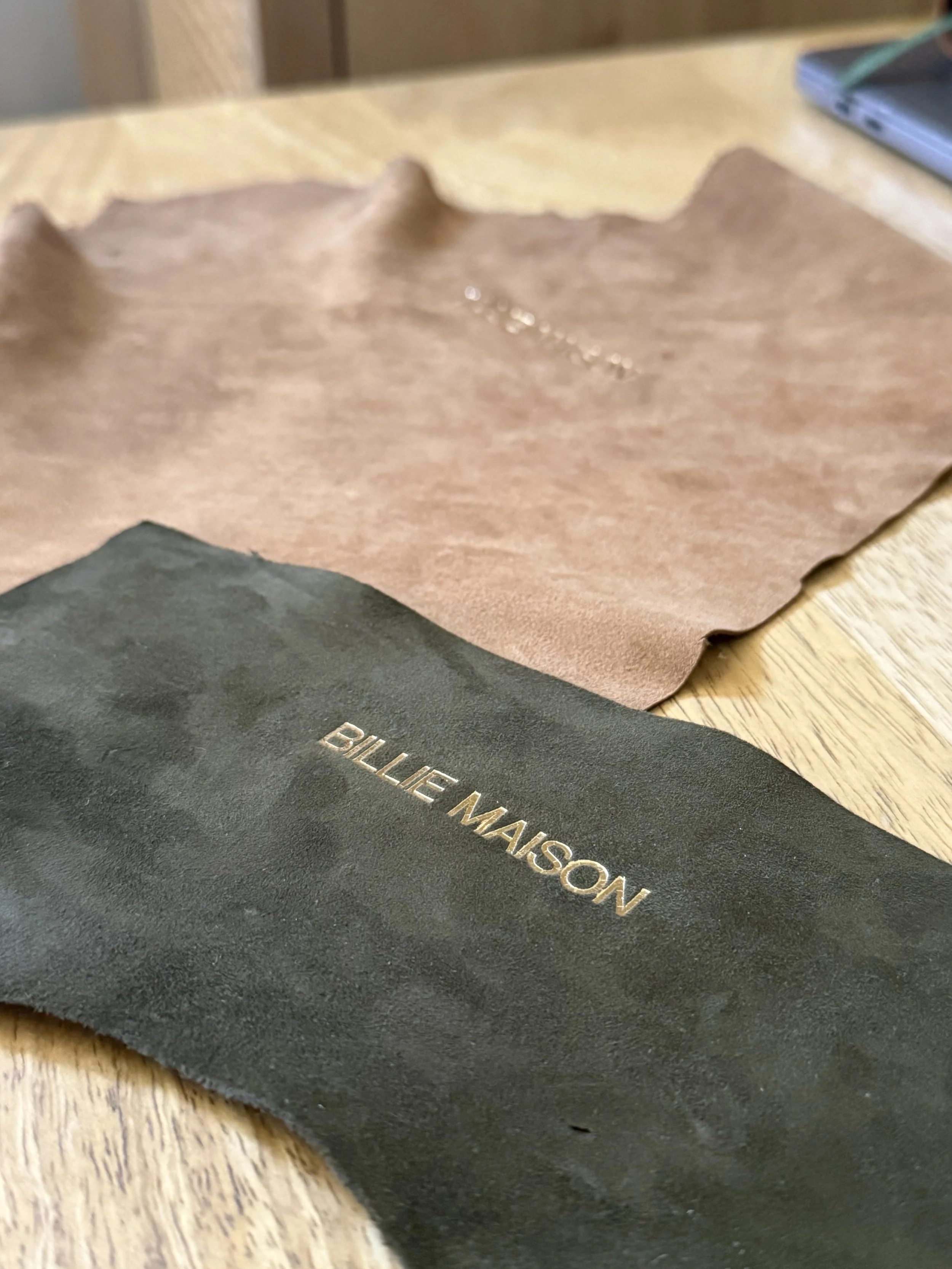 Close-up of black paper with gold embossed text reading 'Billie MAISON' on a wooden table, with brown and gray papers in the background.