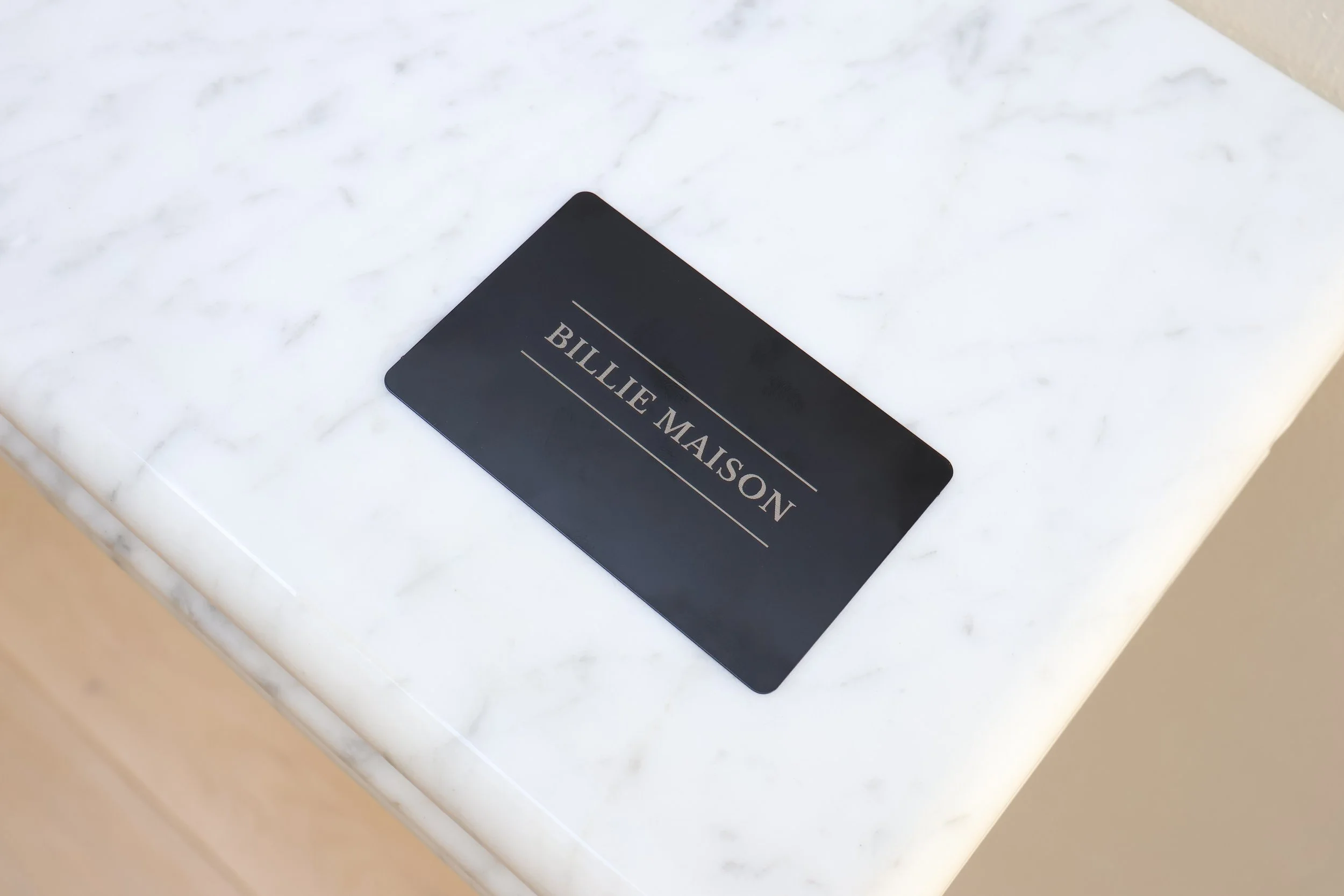 Close-up of a black card with silver text on a white marble surface, with a beige wall in the background.