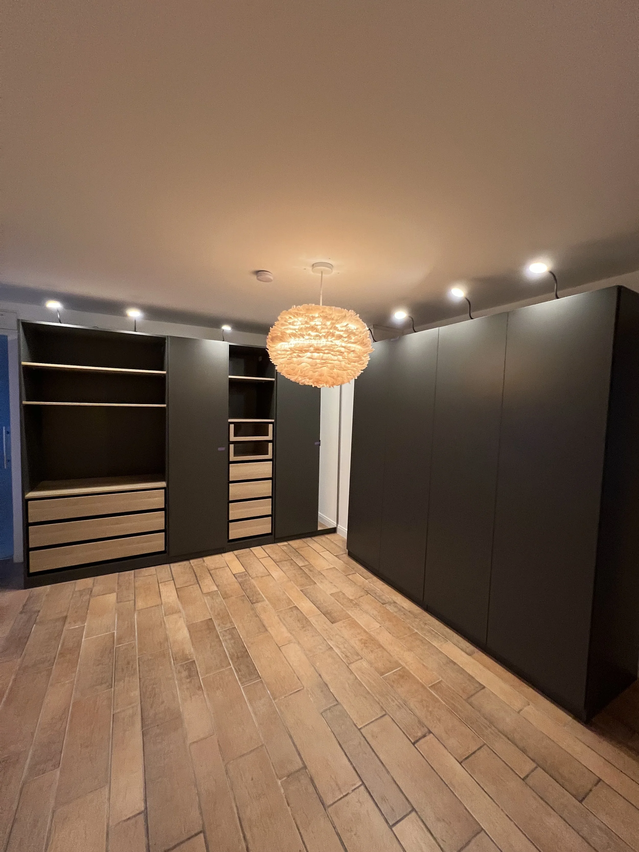 Moderner Raum mit schwarzen Schränken und Holzregalen, Holzboden, zentraler runder Lichtquelle an der Decke.