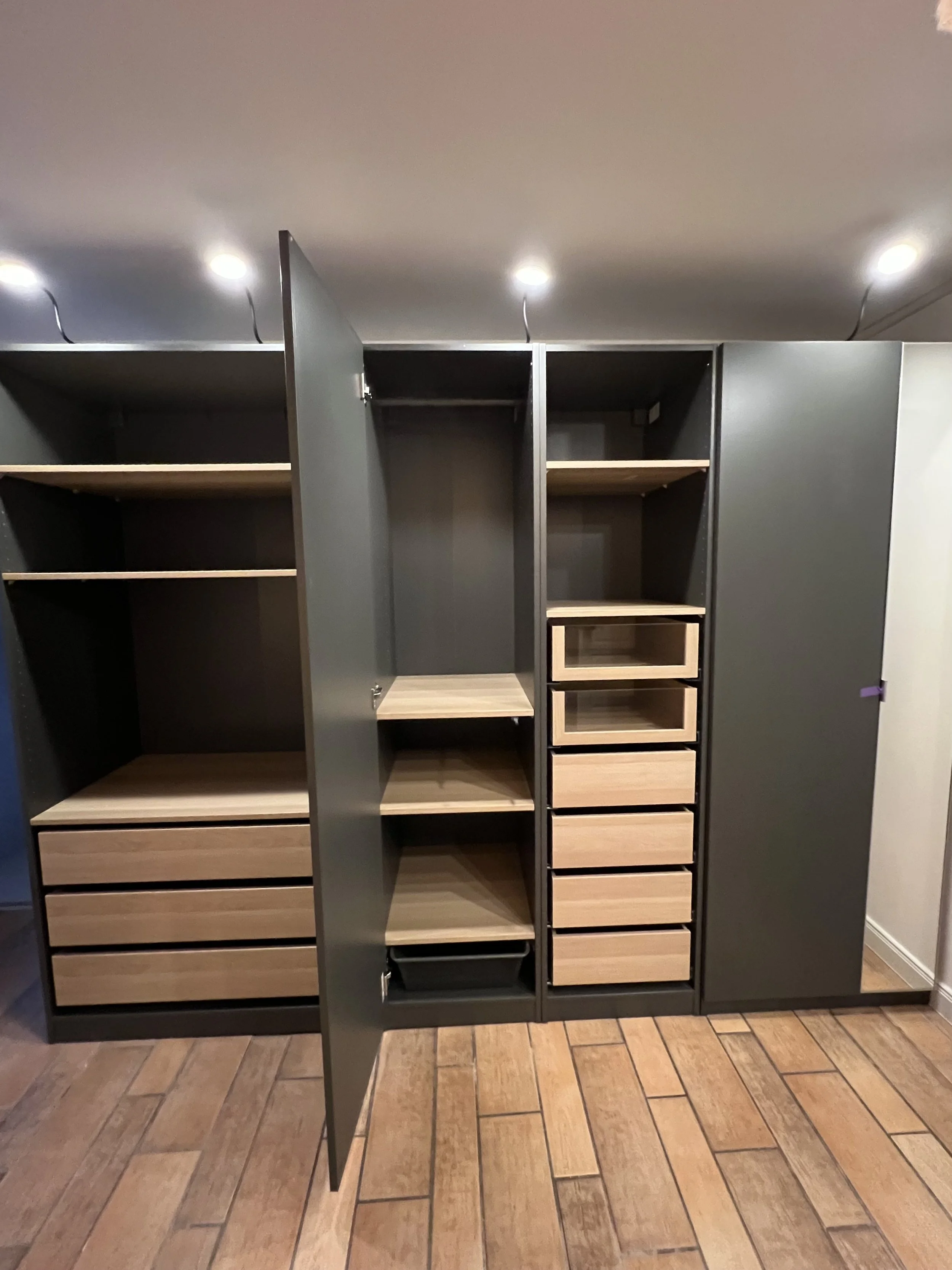 Ein moderner, schwarzer Kleiderschrank mit offenen Türen, bestehend aus mehreren Fächern, Schubladen und offenen Regalen, steht auf einem holzfarbenen Boden.